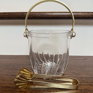 May include: A clear glass ice bucket with a gold-colored handle and matching ice tongs. The bucket has a textured, ridged design. The tongs are resting on a wooden surface.