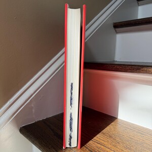 May include: A hardcover book with a bright red spine and edges, standing upright. The pages are white, and there is some dark marking on the page edges. The book is positioned on a wooden surface.