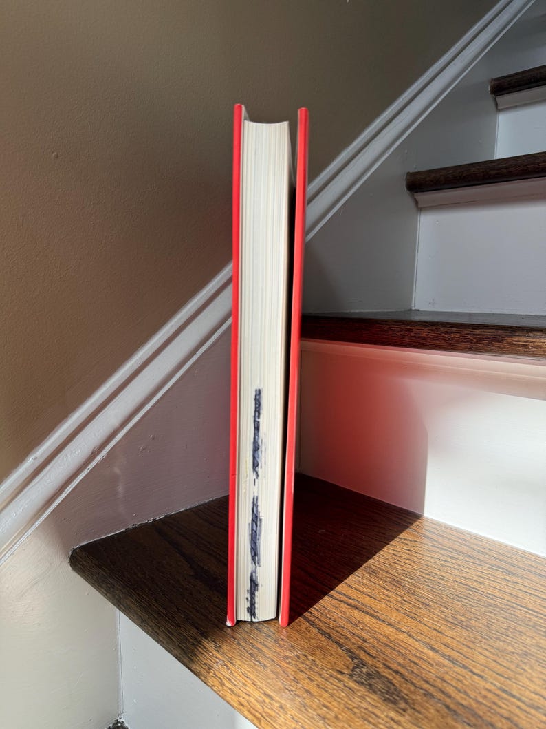May include: A hardcover book with a bright red spine and edges, standing upright. The pages are white, and there is some dark marking on the page edges. The book is positioned on a wooden surface.