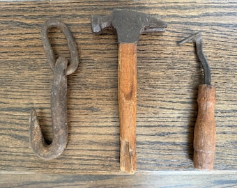 May include: Three antique metal tools on a wooden surface. The tools include a hook, a hammer with a wooden handle, and a curved tool with a wooden handle. The tools are rusty and show signs of wear.