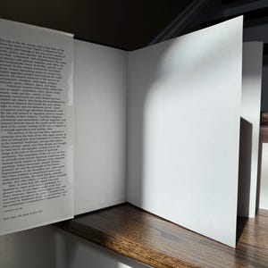May include: An open book with white pages and a red spine, resting on a wooden surface. The left page contains printed text, while the right page is blank. The book is partially open, with the pages angled towards the viewer. The text on the left page is small and dense.