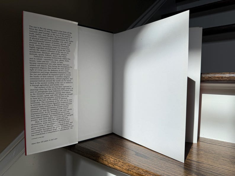 May include: An open book with white pages and a red spine, resting on a wooden surface. The left page contains printed text, while the right page is blank. The book is partially open, with the pages angled towards the viewer. The text on the left page is small and dense.
