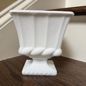 May include: A white milk glass vase featuring a square-shaped top with vertical fluted panels, a decorative rope-like embossed band around the midsection, and a square pedestal base with a matching rope-textured border along the bottom edge.