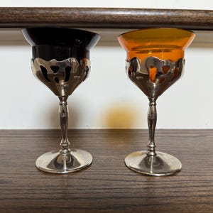 May include: Two antique wine glasses with silver filigree bases and stems. One glass has a black bowl, the other an orange bowl. The glasses are displayed on a dark wooden surface.