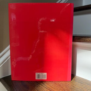 May include: A red book with a glossy cover. The book is standing upright on a wooden surface. A barcode is visible near the bottom of the book.