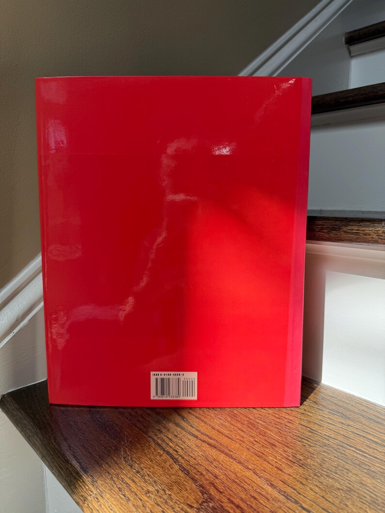 May include: A red book with a glossy cover. The book is standing upright on a wooden surface. A barcode is visible near the bottom of the book.