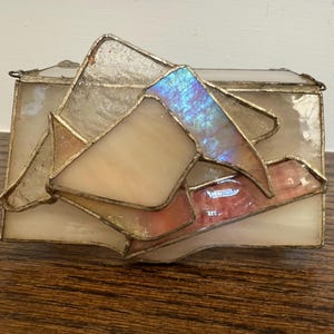 May include: A rectangular stained glass box with a hinged lid. The box features a geometric design with iridescent and pastel-colored glass pieces, framed by a metal border. The colors include cream, pink, and blue.