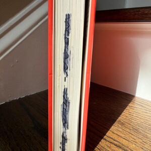 May include: A hardcover book with a bright orange cover and a stack of white pages. The edges of the pages have dark blue markings. The book is resting on a wooden surface.