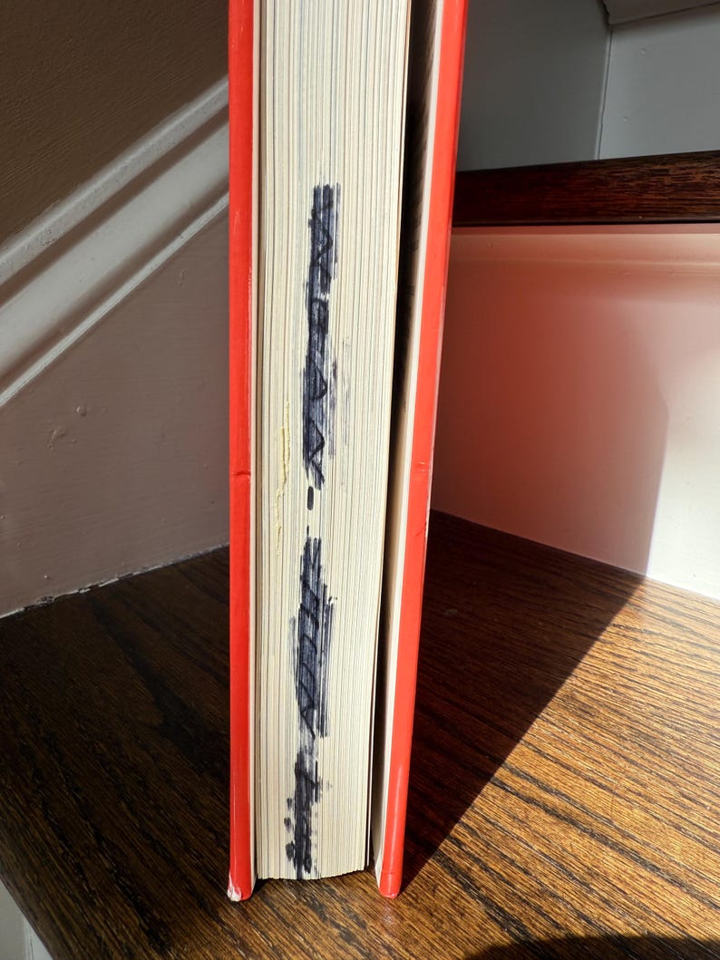 May include: A hardcover book with a bright orange cover and a stack of white pages. The edges of the pages have dark blue markings. The book is resting on a wooden surface.