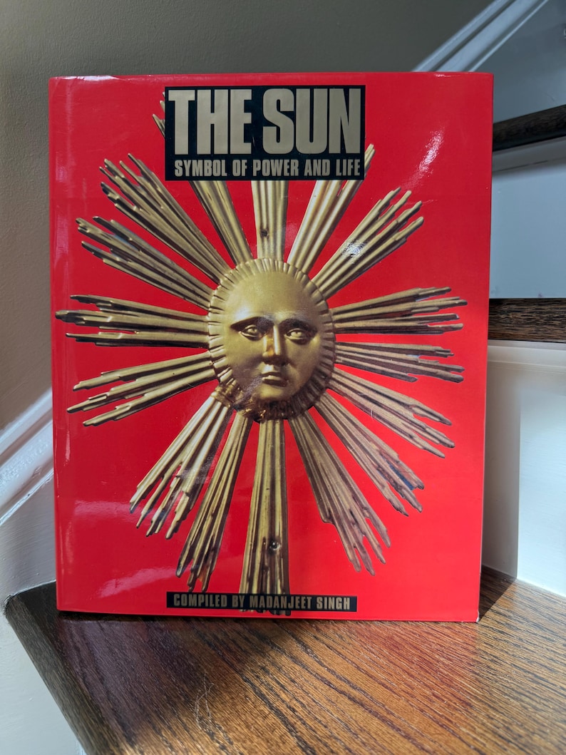 May include: A hardcover book titled "THE SUN" with the subtitle "SYMBOL OF POWER AND LIFE" on a red background. The cover features a golden sun with a face and radiating beams. The author's name, MADANJEET SINGH, is at the bottom.