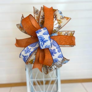 May include: A decorative bow featuring layers of orange, blue, and white ribbons. The bow has a central blue and white floral patterned ribbon, with orange ribbons and ribbons with fall leaf patterns. The bow is placed on top of a white lantern.