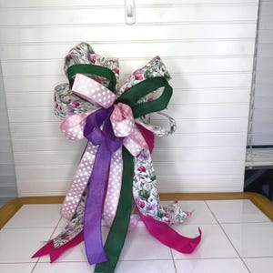 Peut inclure: Un nœud décoratif composé de rubans variés. Le nœud comprend des rubans à pois blancs sur fond rose, des motifs floraux, du violet uni, du vert et du fuchsia. Conçu pour la décoration.