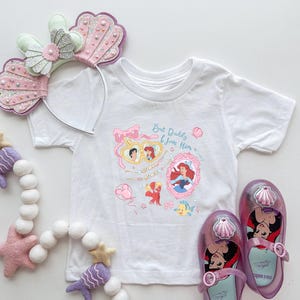 Op de afbeelding: Wit T-shirt met een roze, blauwe en gele afbeelding van Ariel en Prins Eric uit De Kleine Zeemeermin. De afbeelding bevat de tekst "But Daddy I Love Him".