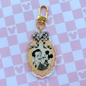 Peut inclure: Un porte-clés en acrylique transparent avec une image en noir et blanc d'un homme et de Mickey Mouse dans un cadre doré. Le porte-clés a un fermoir doré et un nœud à carreaux noir et blanc en haut.