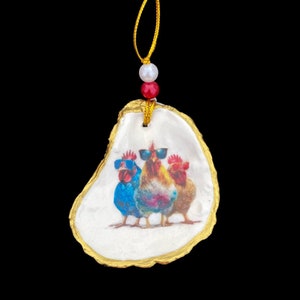 May include: A white oyster shell ornament with a gold trim, featuring a painted image of three roosters wearing sunglasses. The ornament has a red and white bead string for hanging.