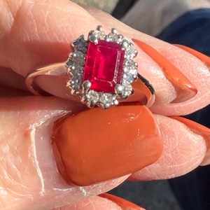 May include: A silver-toned ring featuring a rectangular, emerald-cut red gemstone at the center, surrounded by a halo of small, clear, round-cut stones set in a prong setting on a polished metal band.
