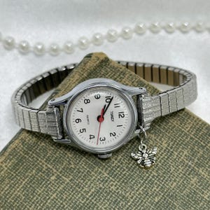 May include: A silver-toned Timex quartz watch with a white face, black numerals, and red second hand. The watch has a stretch band and a small bee charm. The watch is displayed on a textured green surface.