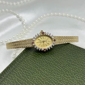 May include: A gold-tone watch with an oval face, a textured woven band, and a bezel set with clear stones and small red accents. The watch dial features the text Mathey-Tissot and Quartz, with gold hands and markers against a gold background.