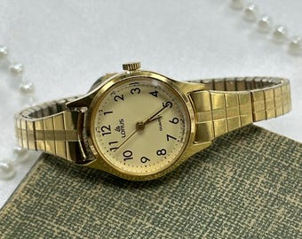 May include: Gold-toned Lorus Quartz wristwatch with a cream-colored face and gold hands and numerals. The watch has a gold expanding band. The watch is resting on a textured green surface.