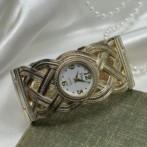 May include: A gold-toned bangle watch with a white face and black hands. The watch face has the word "Viva" and "Quartz" printed on it. The watch band is a woven design.