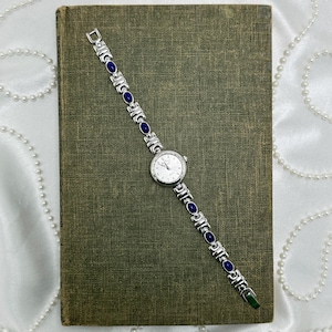 May include: A silver-toned wristwatch with a white face and blue gemstone accents. The watch is displayed on a vintage green book. The watch band features alternating silver links and oval blue stones. The background includes a white fabric and a string of pearls.