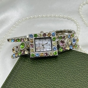 May include: A silver-tone square wristwatch with a white face featuring the text Vivani and Quartz. The bezel and the flexible band are decorated with multi-colored rhinestones in shades of blue, green, pink, and yellow, arranged in rows.
