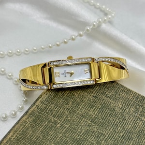 May include: A gold-toned Bulova watch with a rectangular face and a white dial. The watch features diamond accents along the bezel and band. Roman numerals mark the hours. A pearl necklace is draped nearby.