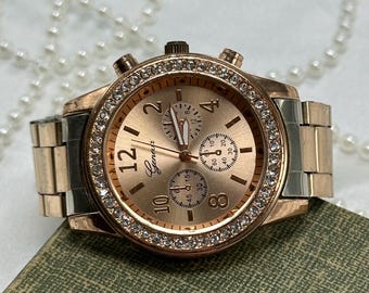May include: A rose gold-toned wristwatch with a crystal-studded bezel and a matching face. The watch features a metallic band with silver and rose gold links. The watch face displays the brand name "Genevr" and has three sub-dials.