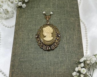 May include: A vintage cameo pendant necklace featuring a cream-colored profile set in an oval, ornate gold-tone frame. The pendant is suspended from a delicate chain with two small pearl accents. The necklace rests on a textured, green-gray book.