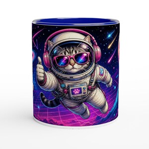 Peut inclure: Un mug en céramique à l'intérieur bleu foncé orné d'un graphique représentant un chat tigré portant une combinaison d'astronaute blanche, un casque audio rose et des lunettes de soleil roses. Le chat fait un pouce levé sur un fond spatial.