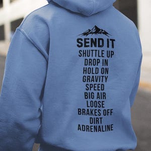 以下が含まれることがあります： 山岳グラフィックと「SEND IT」の文字が入った水色のパーカー。その下に、黒い文字で「SHUTTLE UP, DROP IN, HOLD ON, GRAVITY, SPEED, BIG AIR, LOOSE, BRAKES OFF, DIRT, ADRENALINE」と書かれています。
