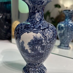 May include: A ceramic vase featuring a blue and white transferware pattern. The design includes a central landscape scene with trees and foliage, surrounded by intricate geometric and floral borders. The vase has a flared rim and a pedestal base.
