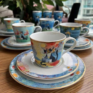 Könnte beinhalten: Ein Porzellan-Teeservice bestehend aus einer Tasse und einer Untertasse mit einer farbenfrohen Illustration von zwei Katzen. Das Design umfasst gelbe, blaue und rote Akzente mit Goldrand an den Kanten. Das Set ist Teil einer passenden Geschirrserie.
