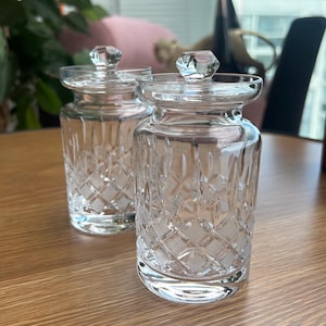 May include: Two clear glass jars with matching lids, each featuring a faceted diamond-shaped finial on top. The cylindrical bodies of the jars are decorated with a cut glass pattern consisting of intersecting lines and small squares.