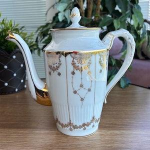 May include: A white porcelain teapot featuring a tall, cylindrical body with vertical fluting at the base. The teapot is decorated with intricate gold filigree patterns, a gold-trimmed spout, a gold-accented handle, and a matching lid with a finial.