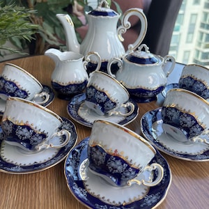 Op de afbeelding: Een theeservies van wit porselein met een diepblauwe band en ingewikkelde gouden filigraanpatronen. De set bevat een theepot met deksel, een melkkan, een suikerpot met deksel en diverse theekopjes met bijpassende schoteltjes met geschulpte randen.