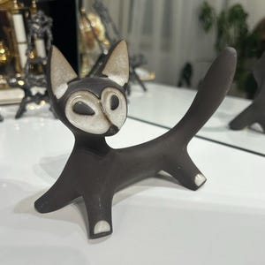 May include: A dark brown ceramic cat figurine with a stylized white face, pointed ears, and a long, upright tail. The cat features small white accents on its paws and a smooth, matte finish, standing on four legs in a poised, alert stance.