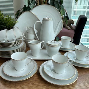 May include: A white porcelain tea set featuring a tall teapot, a lidded sugar bowl, a creamer, multiple cups, saucers, and plates. Each piece is decorated with a delicate gold-toned geometric pattern along the rims and edges of the white ceramic.