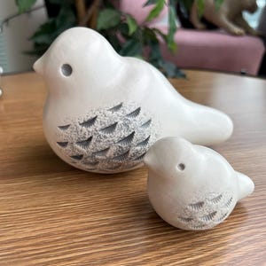 Könnte beinhalten: Zwei cremeweiße Keramikvogel-Figuren in verschiedenen Größen. Jeder Vogel hat einen glatten, abgerundeten Körper mit einem kleinen kreisförmigen Auge und einem texturierten Flügelbereich, der mit kleinen, dunklen, V-förmigen Einkerbungen verziert ist.