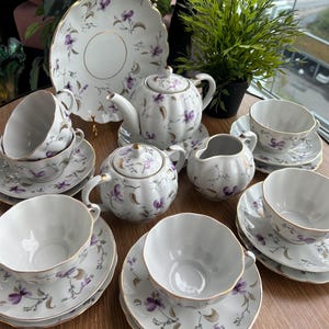 Puede incluir: Un juego de té de porcelana blanca con un delicado patrón floral de violetas moradas y bordes dorados. El conjunto incluye una tetera, un azucarero con tapa, una lechera, varias tazas con sus platos y un plato grande con bordes festoneados.