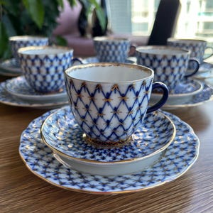 Puede incluir: Un juego de tazas de té y platillos de porcelana con base blanca, patrón de rejilla azul y detalles dorados. Cada taza tiene una pequeña asa azul oscuro y un borde dorado, a juego con los bordes festoneados de los platillos circulares.