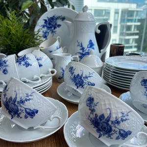 Puede incluir: Un juego de té de porcelana blanca con un patrón floral azul y textura de celosía en relieve. La colección incluye varias tazas con sus platos, una tetera con tapa, una jarra para leche, un azucarero y varios platos llanos apilados.