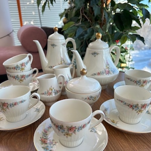 Könnte beinhalten: Ein Teeservice aus Porzellan mit Kaffeekanne, Teekanne, Zuckerdose, zwei Milchkännchen und sechs Tassen mit passenden Untertassen. Jedes Teil ist cremefarben mit einem zarten Blumenmuster in Blau, Rosa und Gelb sowie goldenen Randverzierungen.