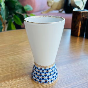 May include: A white porcelain tumbler with a flared rim and a tapered base. The rim features a thin gold band with a delicate scalloped pattern. The base is decorated with a blue and gold lattice design featuring small cross-shaped accents.