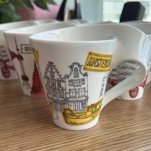Może przedstawiać: Biały ceramiczny kubek z unikalnie zakrzywionym uchwytem, ozdobiony czarnymi rysunkami liniowymi amsterdamskich kamienic, żółtą łodzią taksówkową i napisem AMSTERDAM w żółtej ramce, z żółtymi akcentami oraz ilustracją tulipana.