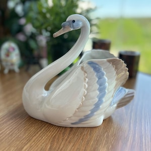 Peut inclure: Une figurine de cygne en céramique blanche avec un long cou courbé et un centre creux. Le cygne présente des accents délicats bleu clair et beige sur ses ailes et sa tête, avec une finition lisse et brillante et un motif de plumes sculpté.