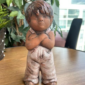 Könnte beinhalten: Eine Terrakotta-Figur eines Kindes mit dunkelbraunem Kopf und Haar, das eine hellfarbene, verwittert wirkende Latzhose trägt. Die Figur ist mit vor der Brust verschränkten Armen, einem neutralen Gesichtsausdruck und einer matten Oberfläche dargestellt.
