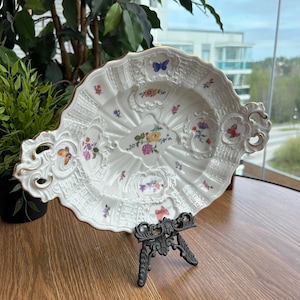 Może przedstawiać: Owalny półmisek z białej porcelany ze złoconymi, falistymi brzegami i uchwytami. Powierzchnia ozdobiona jest tłoczonym wzorem plecionki, kolorowymi bukietami kwiatów oraz drobnymi motywami motyli rozmieszczonymi na środku i na obrzeżach.
