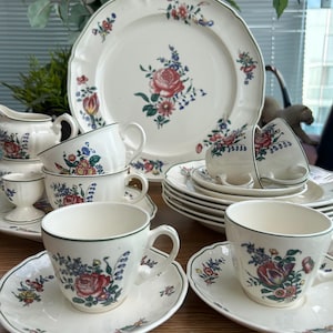Op de afbeelding: Een crèmekleurige keramische serviesset met een delicaat bloemmotief van rode rozen, blauwe bloemen en groene bladeren. De collectie bevat een groot serveerbord, meerdere theekopjes met bijpassende schoteltjes en een kleine melkkan.