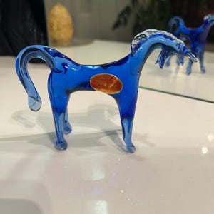 Peut inclure: Une figurine de cheval en verre bleu vif dotée d'une crinière et d'une queue fluides, représentée dans une posture élégante. Un autocollant doré ovale sur le côté porte l'inscription Made in Murano Italy, indiquant son origine et sa fabrication.